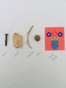 A piece of white paper with labelled objects lined up in a row along the centre: an iron nail, wood chip, knotted leather string, round brown stone, and pink card stock with a print of three flowers in a vase.