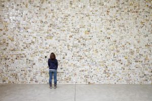 A person stands looking at a wall of photos salvaged from the Japanese earthquake of 2011. The photos are mostly white and yellow-toned.