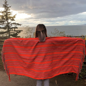 A person facing away from the camera, looking at the ocean. They hold a red shawl across their back with outstretched arms.