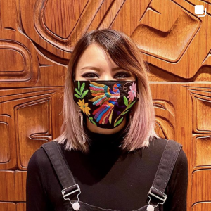 Photo of a person with brown and pink shoulder-length hair, wearing a colourfully embroidered face mask.