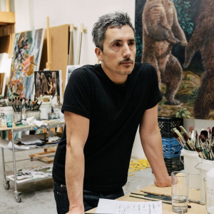 Artist Kent Monkman (Swampy Cree) stands in an art studio. He has short grey hair and a moustache. He wears a black T-Shirt.
