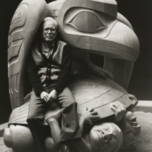 Black and white photograph of Haida artist Bill Reid sitting under the wing of his carving, Raven and the First Men.