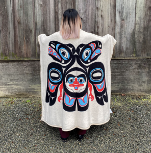 A person wraps a cream blanket around their back. It has a Northwest Coast design of an eagle and a raven, facing outwards.